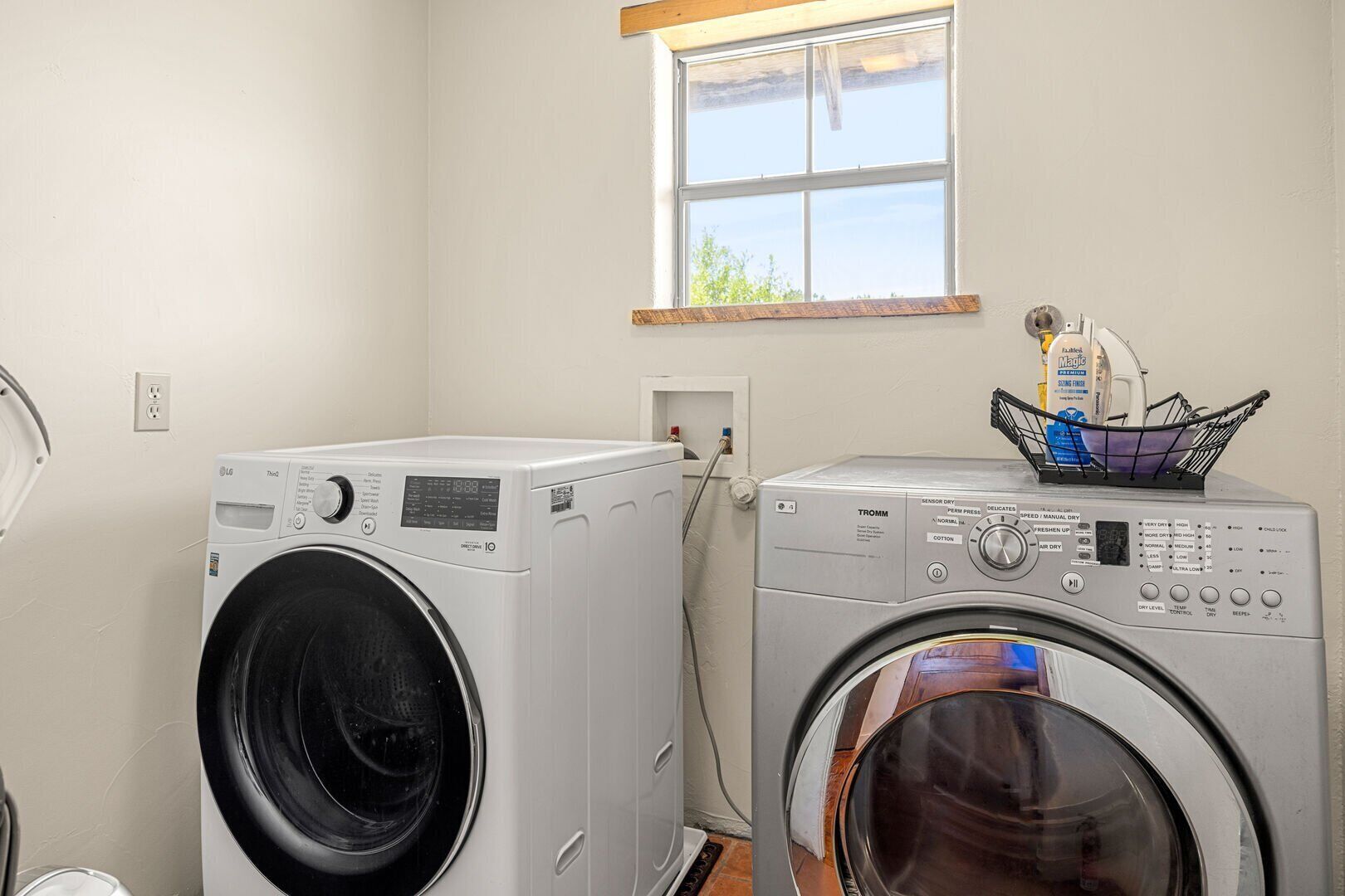 A laundry room with two front-loading washing machines, a window, and a basket with laundry detergent on the wall.