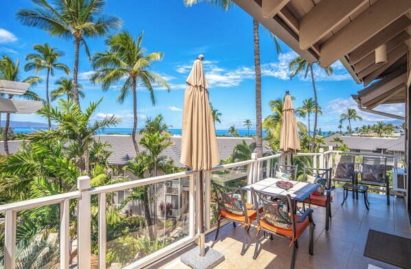 A balcony with a table and chairs, overlooking a tropical landscape with palm trees and a view of the ocean.