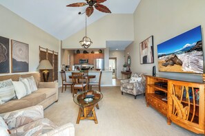 A spacious living room with a sofa, coffee table, and TV. There is a kitchen in the background with wooden cabinets and a dining area.