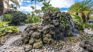Une formation rocheuse naturelle avec une cascade, entourée d’une végétation tropicale et bordée d’un sentier pavé.