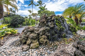 Formazione rocciosa naturale con una cascata, circondata da vegetazione tropicale e un sentiero lastricato.