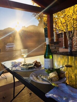 A table with a bottle of wine, glasses, and food items on a glass table with a view of a sunset and a stone building with trees.
