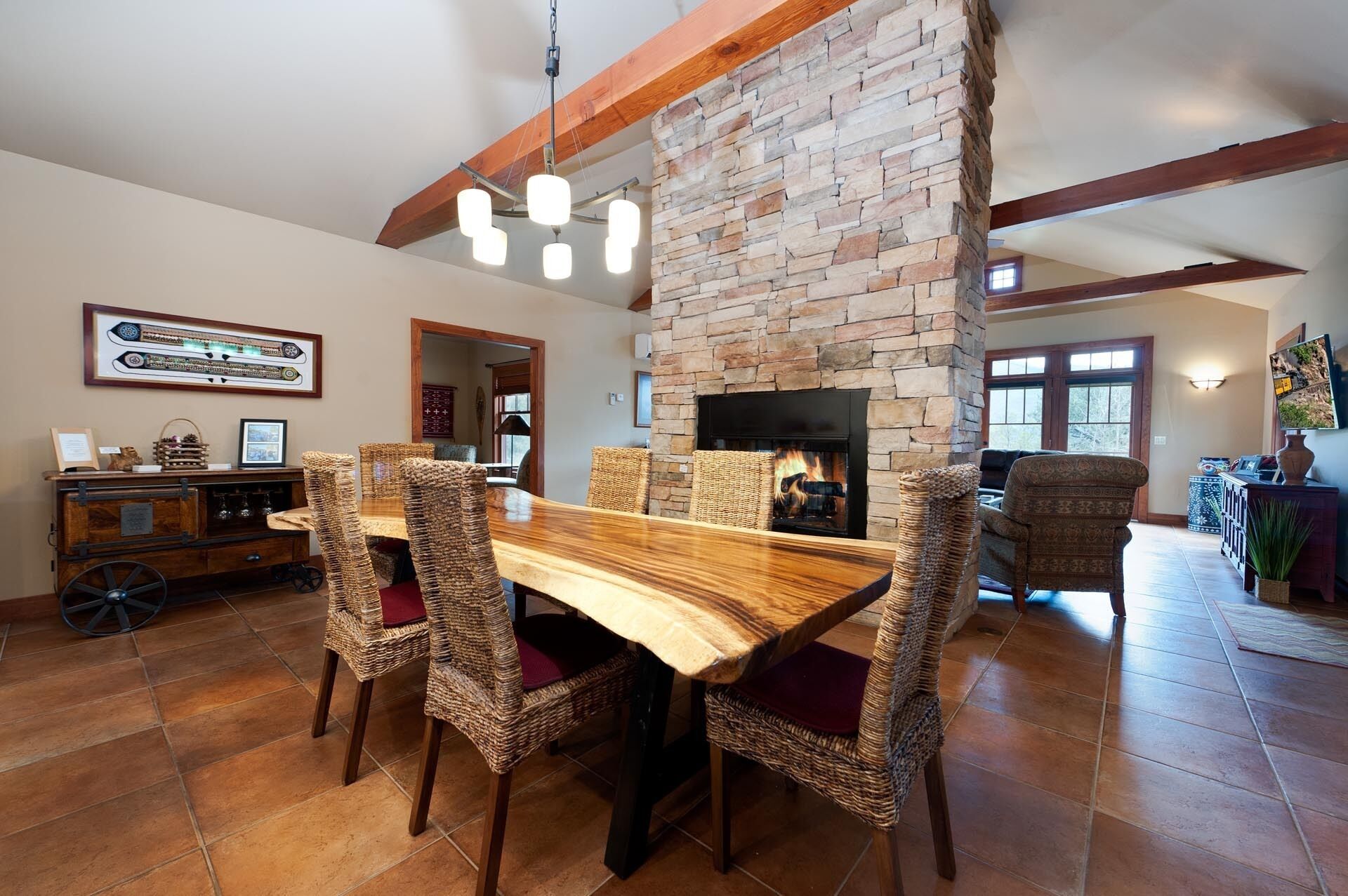 A dining area with a wooden table and wicker chairs, a stone fireplace, and a view of the living room.