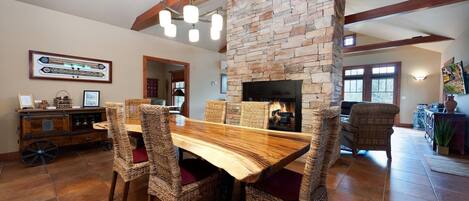 A dining area with a wooden table and wicker chairs, a stone fireplace, and a view of the living room.
