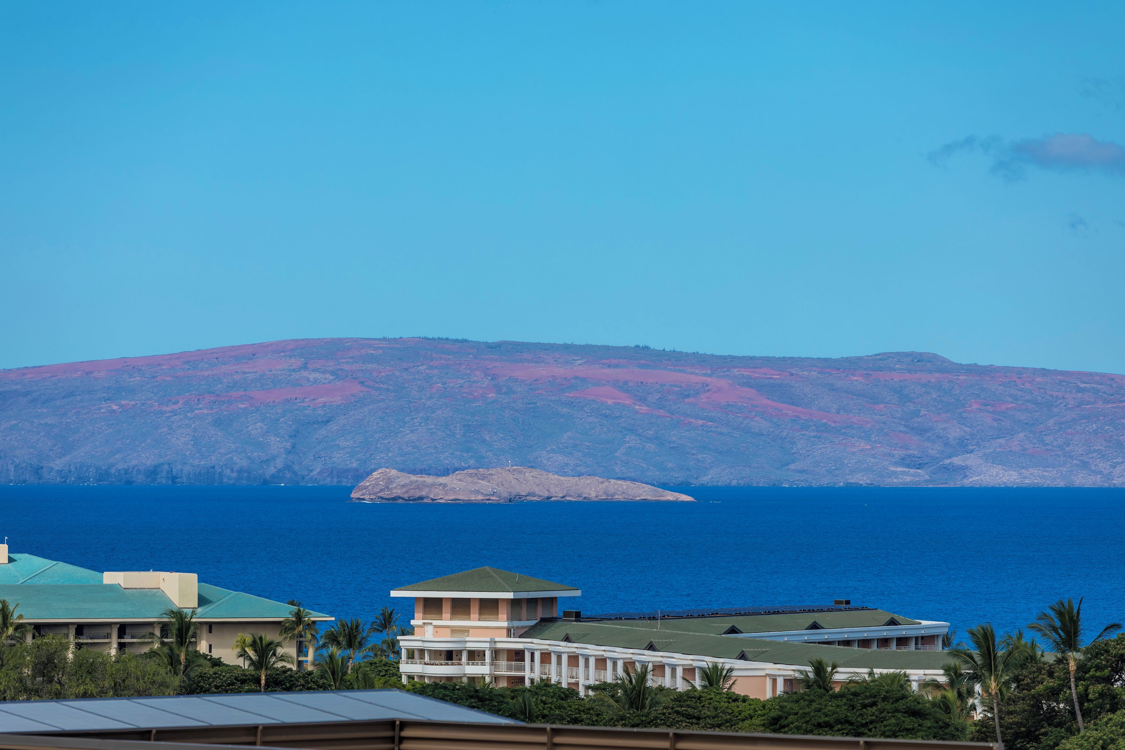 A coastal view with a mountain in the background, a body of water, and buildings with a green roof.