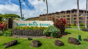 A sign for Lawai Beach Resort with a stone wall, tropical plants, and a multi-story building in the background.