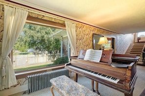 Um quarto com um piano de cauda, um banquinho, um radiador, uma janela com cortinas e uma escadaria.