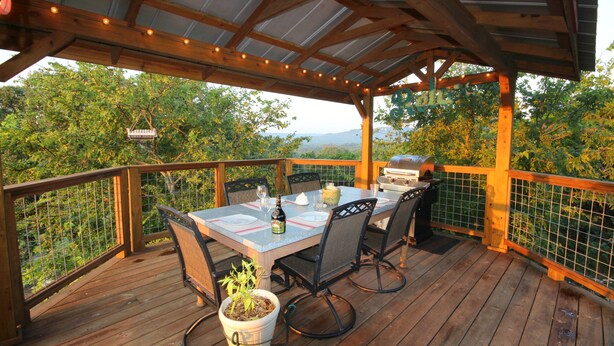 An outdoor wooden deck with a table set for dining, surrounded by chairs, and a grill.