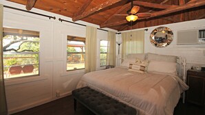 A bedroom with a wooden ceiling, a bed with a white comforter, a round mirror, and a window with curtains.
