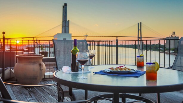 A table with drinks and a clay pot on a balcony overlooking a sunset and a bridge.