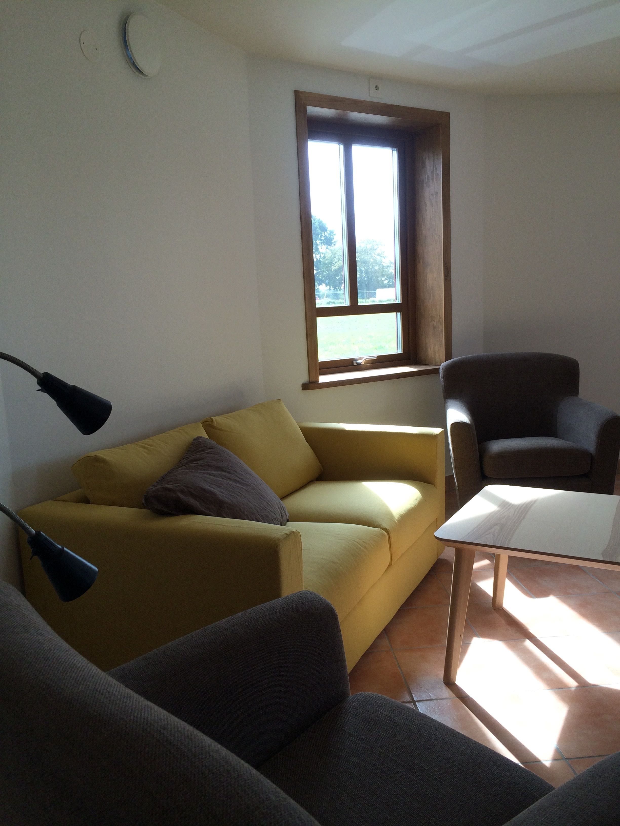 Una sala de estar con un sofá amarillo, un sillón oscuro y una mesita. Se ve una ventana con vistas a árboles y una puerta al fondo.