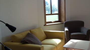 Una sala de estar con un sofá amarillo, un sillón oscuro y una mesita. Se ve una ventana con vistas a árboles y una puerta al fondo.