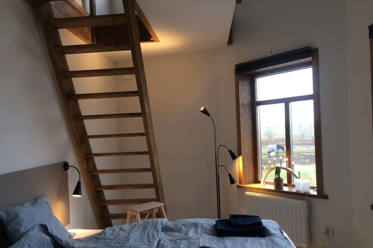 A bedroom with a sloped ceiling, a wooden ladder, a bed with a striped blanket, a small stool, and a window with a view of the outdoors.