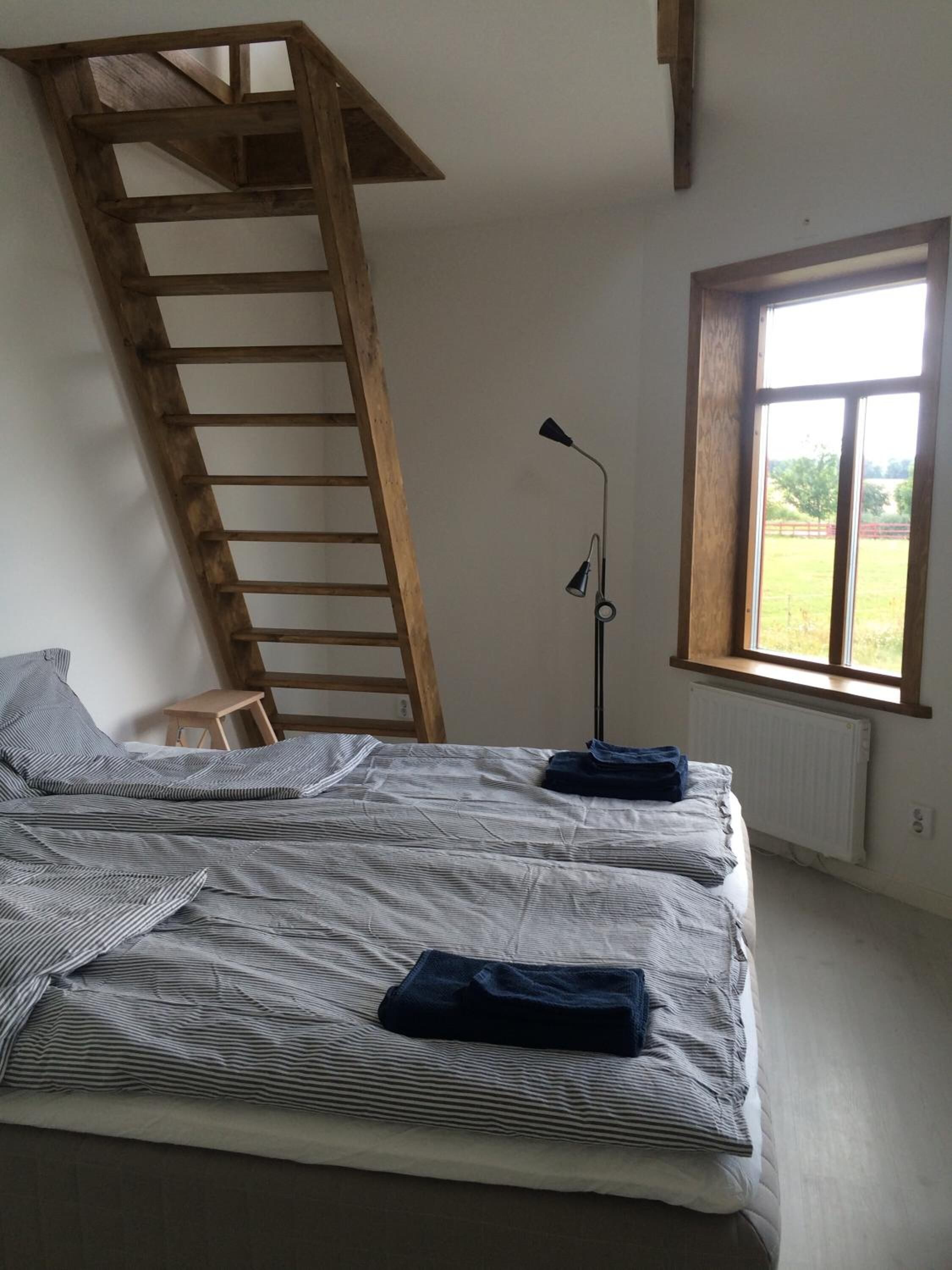 A bedroom with a sloped ceiling, a wooden ladder, a bed with striped bedding, a floor lamp, and a window with a view of a green landscape.