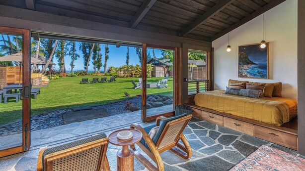 A spacious room with a large sliding glass door leading to a backyard. The room features a bed, a seating area with chairs, and a small table. - Luxurious Beachfront Retreat (Hanalei)