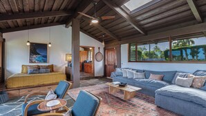 A spacious living area with a large sofa, a coffee table, and a dining table. There is a kitchen in the background with wooden cabinets and a hanging light fixture. - Luxurious Beachfront Retreat (Hanalei)