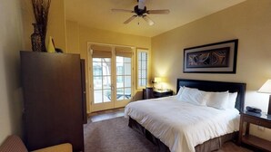 Une chambre d’hôtel avec un lit, un ventilateur de plafond, une table de chevet avec une lampe, un tableau encadré au mur et une porte donnant sur un balcon.