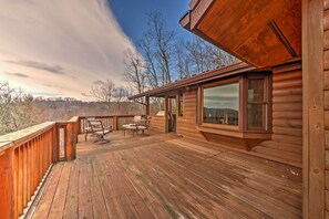 Una terraza de madera con mobiliario exterior, un ventanal y vistas a árboles y montañas.
