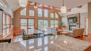 A spacious living area with a large dining table, multiple windows, a stone fireplace, and a ceiling fan.