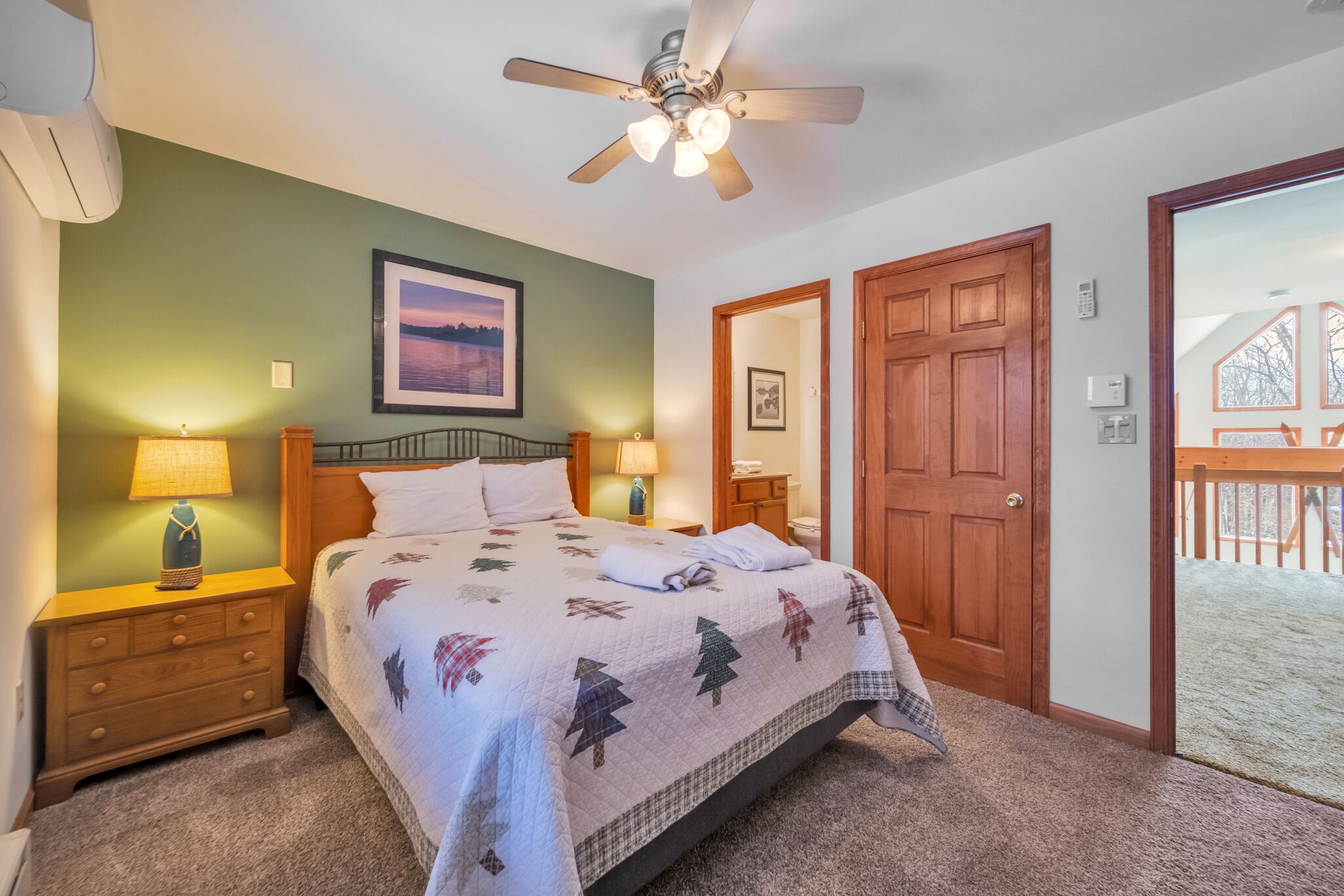 A bedroom with a bed, two bedside lamps, a ceiling fan, a framed picture on the wall, and a wooden door leading to a balcony.