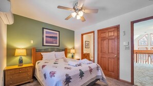 Ein Schlafzimmer mit einem Bett, zwei Nachttischlampen, einem Deckenventilator, einem gerahmten Bild an der Wand und einer Holztür, die auf einen Balkon führt.