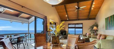 A spacious living area with a glass table, wicker chairs, a sofa, and a television. There is a large window offering a view of the ocean.