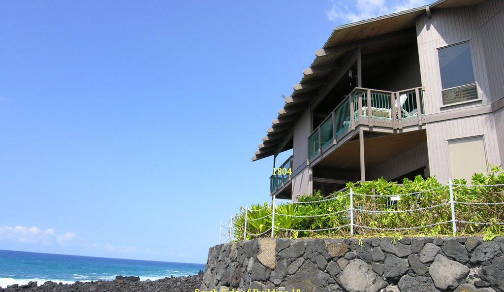 A two-story building with a stone base, a balcony with green railings, and a clear view of the ocean.