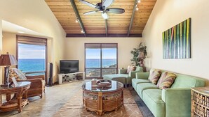 Un salón espacioso con un ventanal grande con vistas al océano, un ventilador de techo, un sofá verde y un televisor.