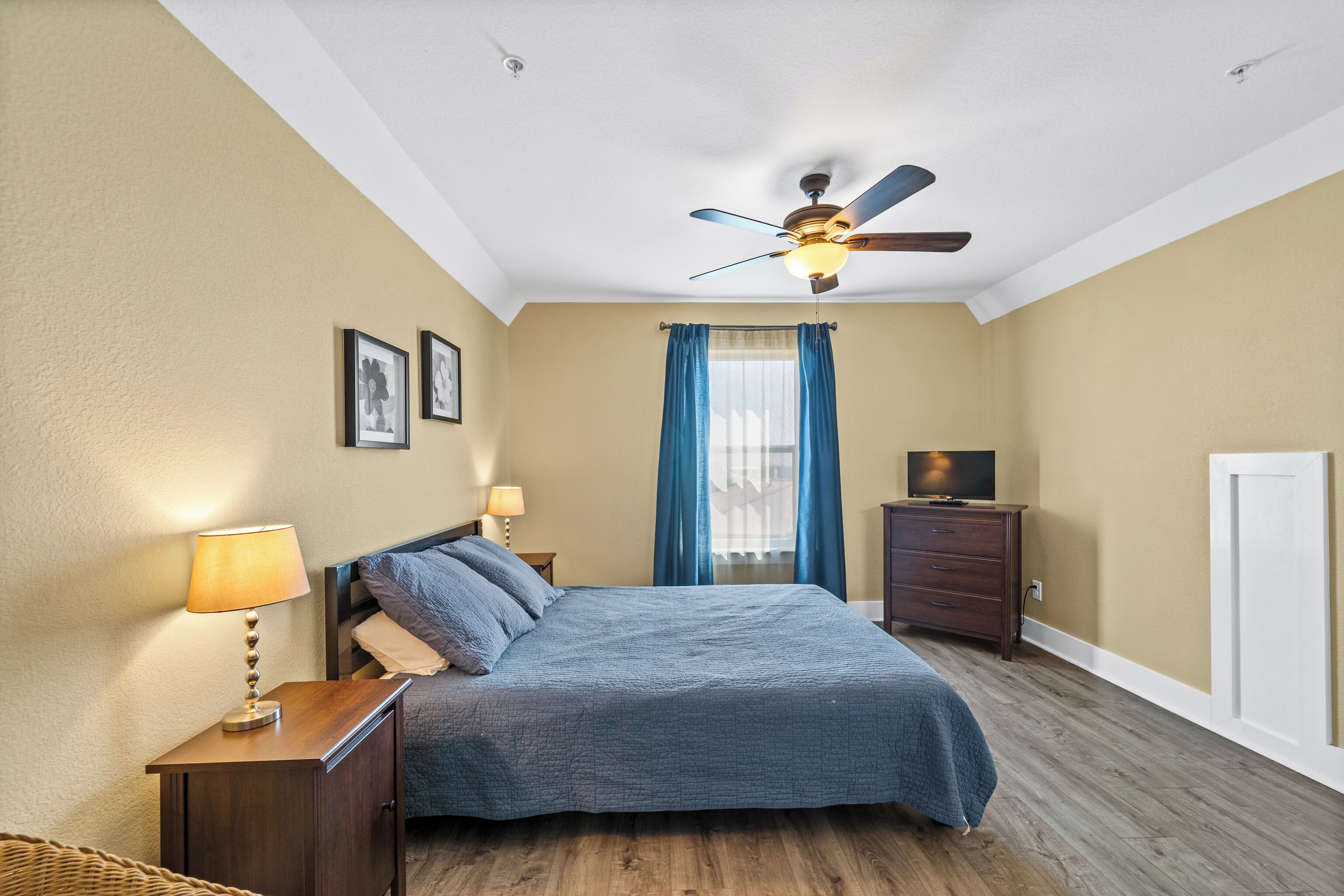 Une chambre à coucher comprenant un lit, un ventilateur de plafond, une table de chevet avec une lampe, une commode et un téléviseur.