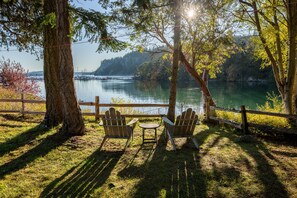 A lakeside seating area with Adirondack chairs and a small table, surrounded by trees and a wooden fence. - Orcas Island, Marina View #201 (Eastsound)