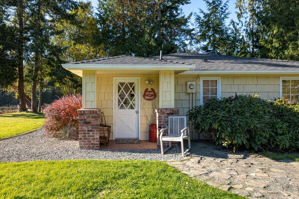 A single-story house with a front porch, a door with a window, and a small brick structure to the left. - Orcas Island, Marina View #201 (Eastsound)