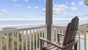 Une chaise en bois sur un balcon donnant sur une plage, avec un ciel dégagé.