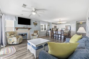 A spacious living room with a fireplace, a dining area with a kitchen in the background, and a ceiling fan.