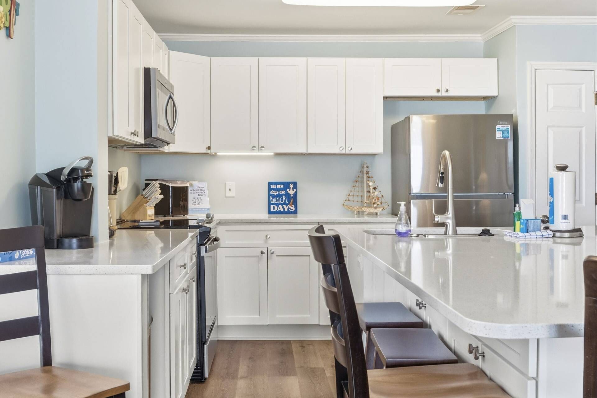 A modern kitchen with white cabinets, a stainless steel refrigerator, a coffee machine, and a sign that reads "The best days are beach days".