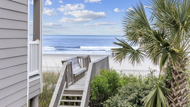 Une terrasse en bois menant à une plage avec vue sur l’océan, un palmier et un bâtiment avec un revêtement extérieur.