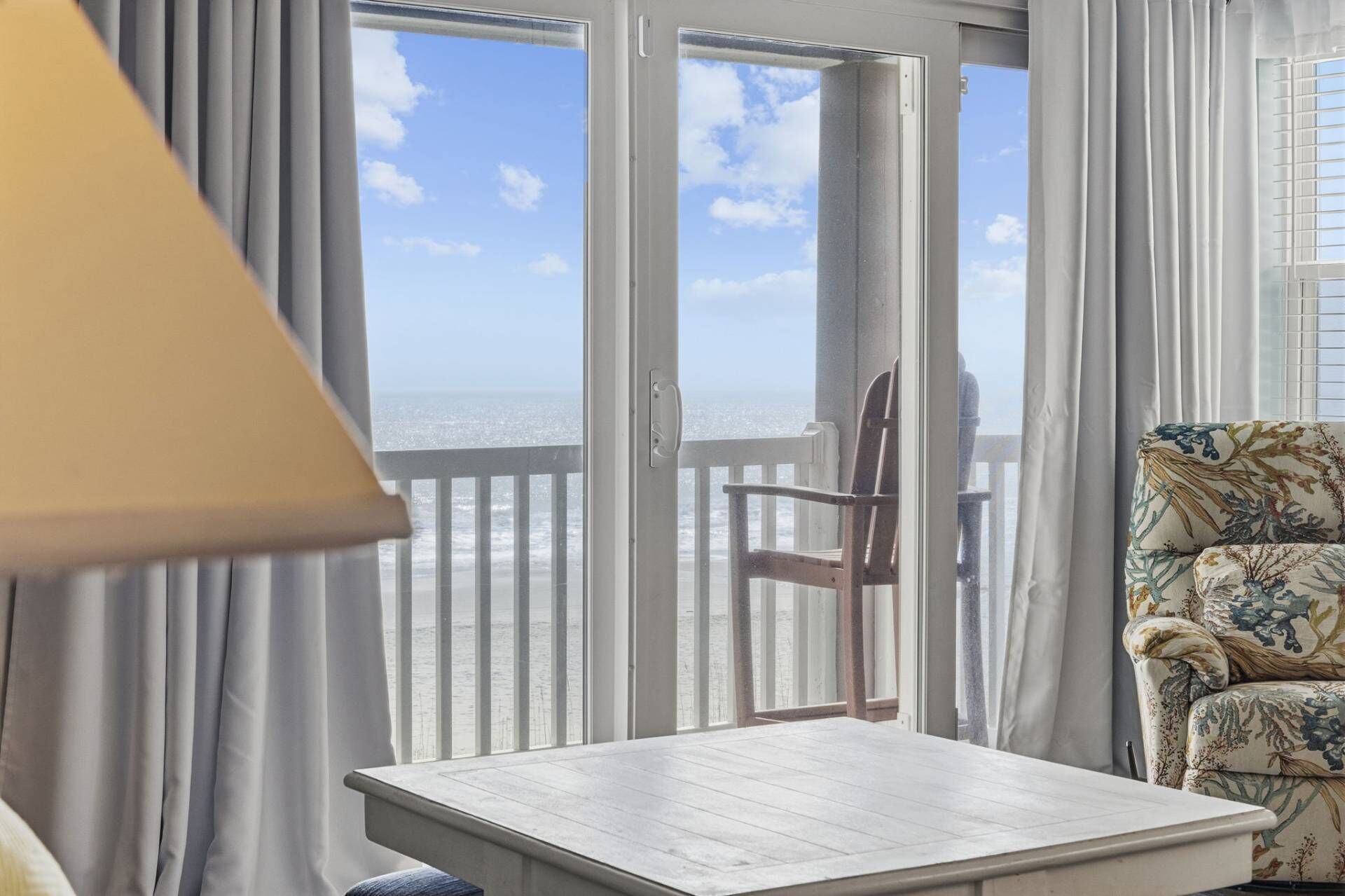 Quarto com vista para o oceano através de uma porta deslizante, uma cadeira de madeira, uma poltrona estampada e uma mesa pequena.
