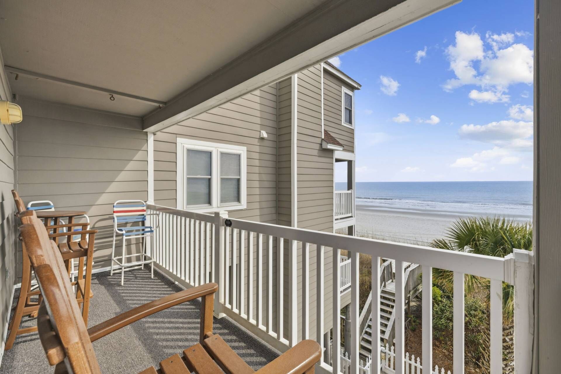 Ein Balkon mit Blick auf den Strand, Holzstühle, ein kleiner Tisch und ein weißes Geländer.