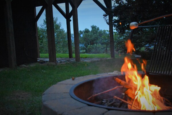 A fire pit with flames, surrounded by a stone structure, under a wooden pergola with a view of a grassy area and trees.