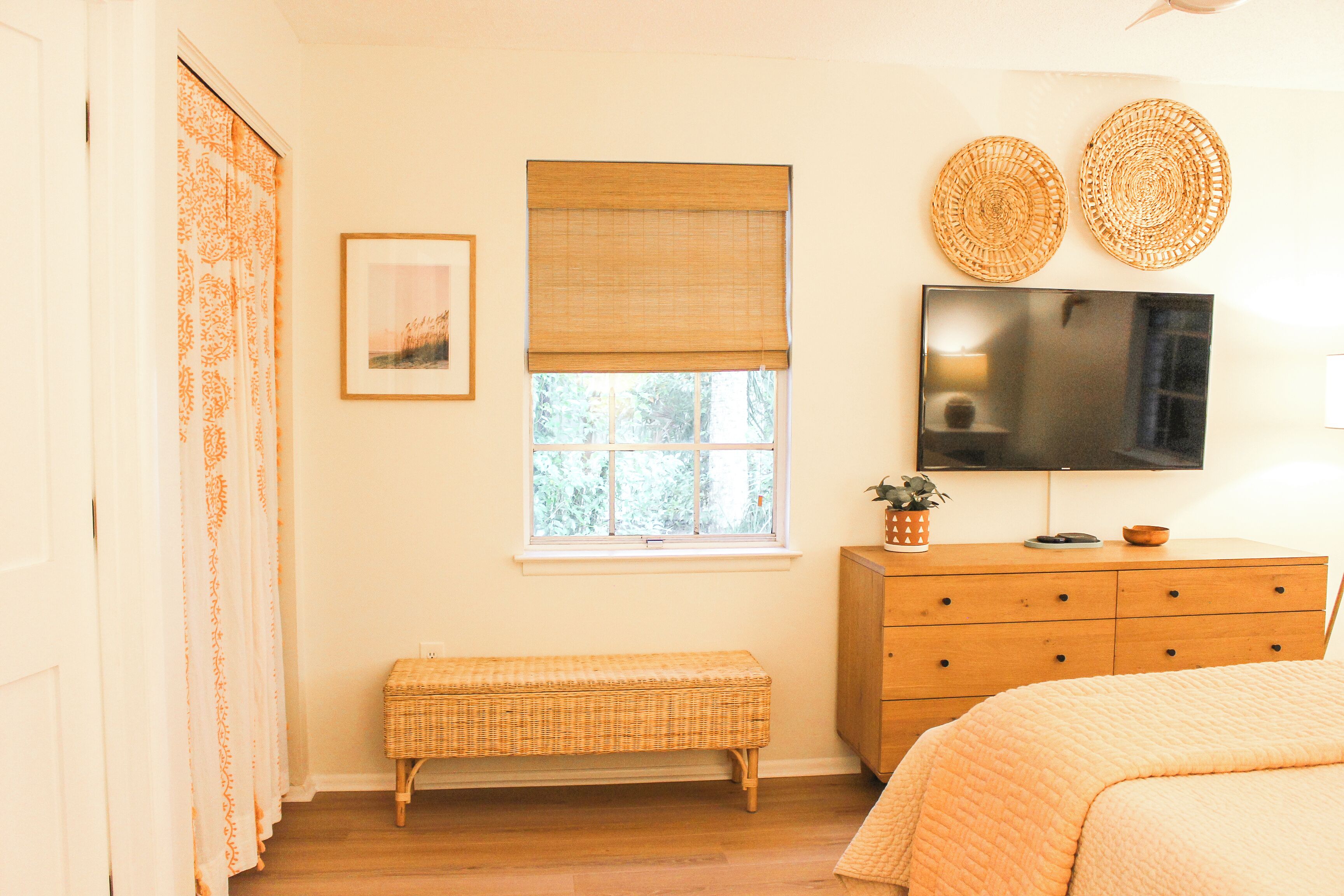 Quarto com cômoda de madeira, televisão, um banquinho e janela com persianas.