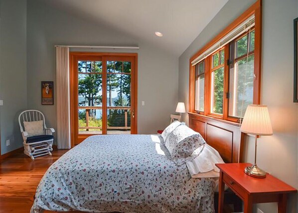 A bedroom with a large bed, a chair, a nightstand, and a window with a view of trees. - Lopez Island, Raven Rock #318 (Lopez Island)