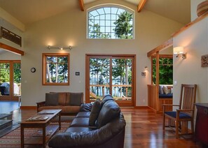 A spacious living room with a leather sofa, wooden coffee table, and a large window offering a view of trees and a deck.