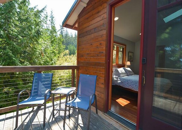 A wooden cabin with a balcony, two blue chairs, and a bed with a patterned blanket.