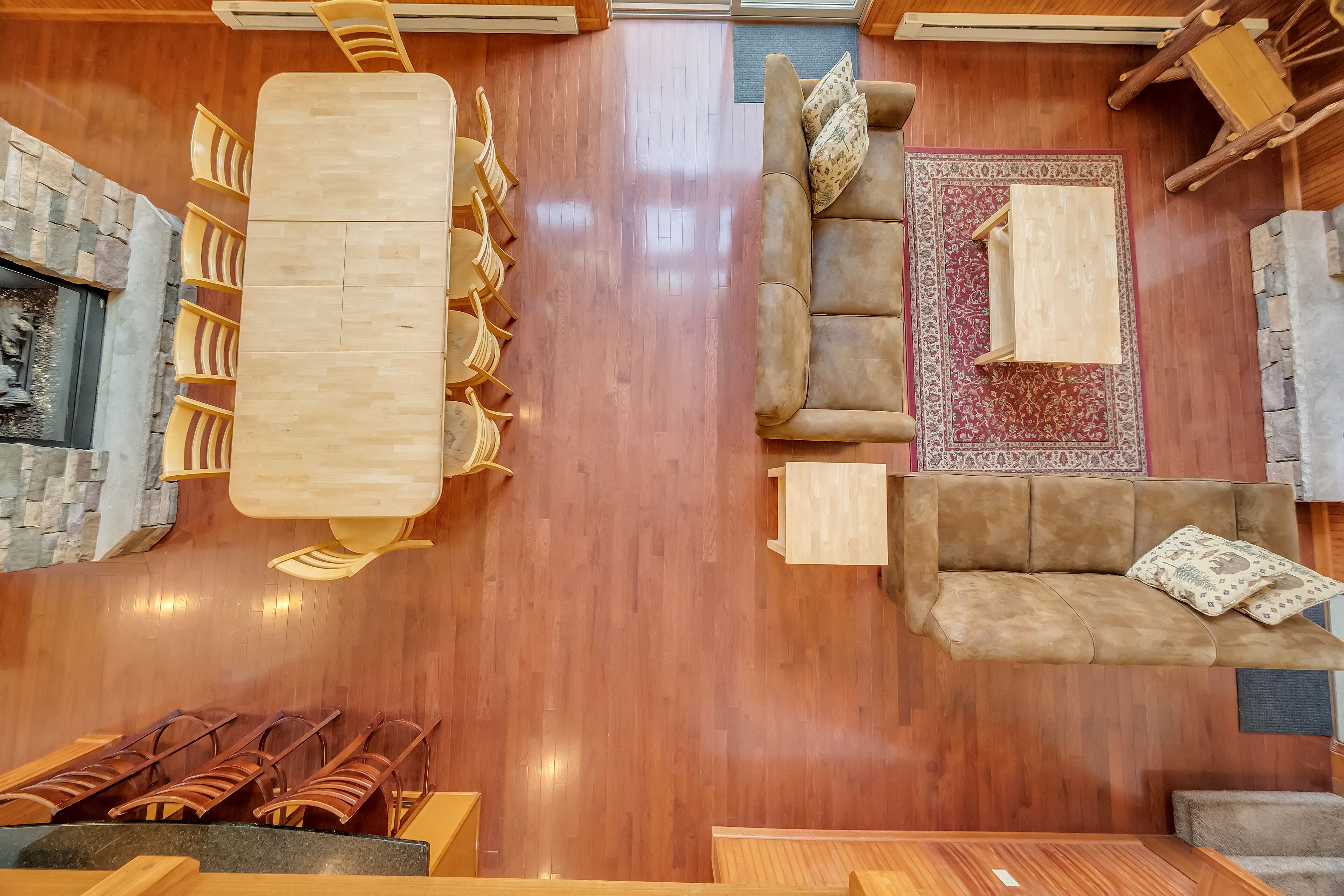 Una habitación amplia con piso de madera, chimenea de piedra, sofá color beige con cojines estampados, alfombra roja y blanca, y mesa de comedor de madera con sillas.