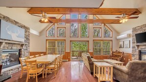 Un vaste espace de vie comprenant une table à manger en bois, des sièges confortables, une cheminée en pierre et de grandes fenêtres offrant une vue sur l’extérieur.