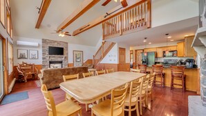 Un vaste espace de vie comprenant une grande table à manger en bois, des sièges confortables, une cheminée et un coin cuisine doté de meubles en bois et d’un mur d’accent en pierre.