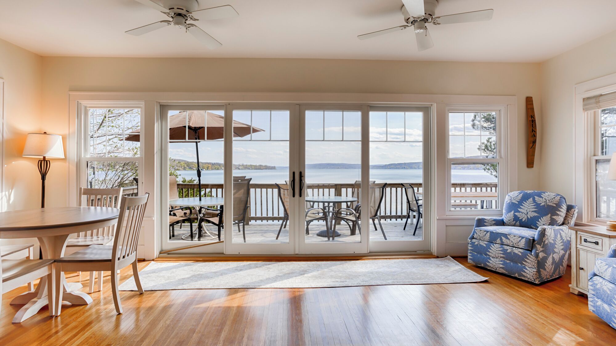 A spacious room with a wooden floor, a dining table with chairs, a blue patterned armchair, and a view of a lake through large sliding glass doors.