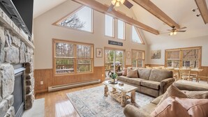 A spacious living room with a stone fireplace, large windows, a high ceiling with wooden beams, and a cozy seating area.