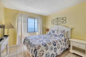 Un dormitorio con cama, mesita de noche, lámpara, ventana con cortinas y vista al mar.