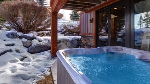 Un jacuzzi con revestimiento de piedra, rodeado de nieve y rocas, con una pérgola de madera y una puerta de cristal que da a un paisaje nevado.