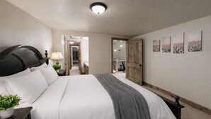 Un dormitorio con una cama grande, una mesita de noche con una lámpara, una planta en maceta y una puerta que da a otra habitación.